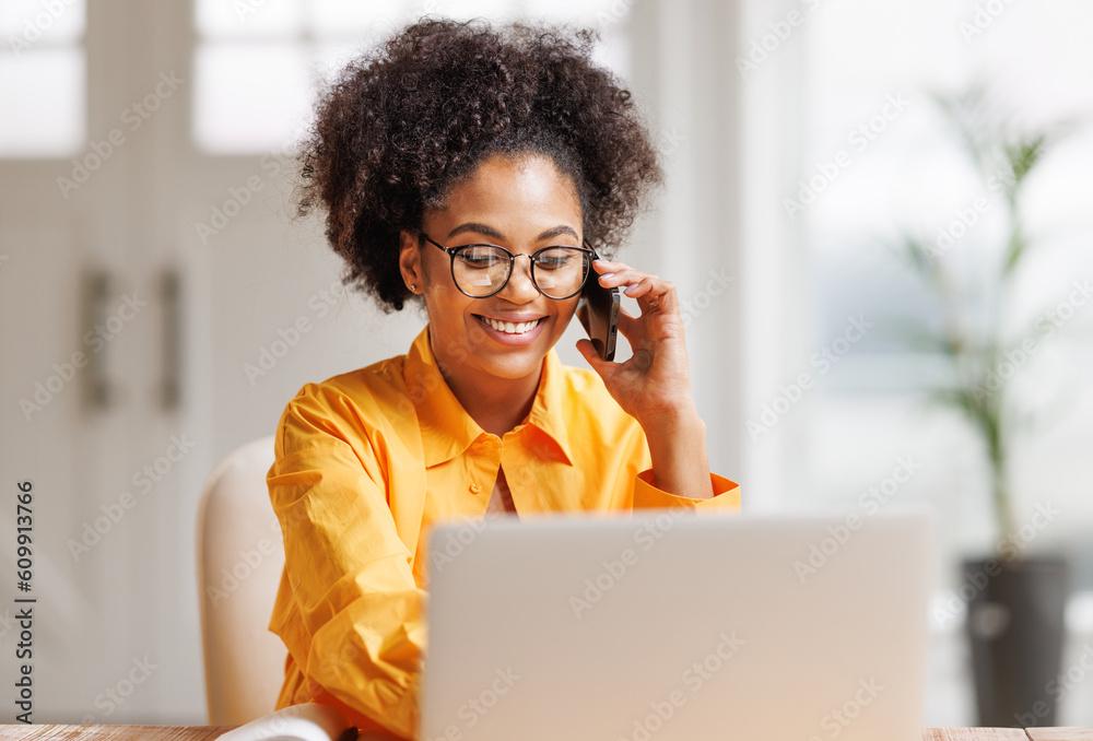 Beautiful young smiling ethnic woman making call via smartphone while working remotely from home while sitting at desk and talking to coworkers .